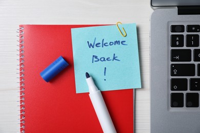 Photo of Paper note with phrase Welcome Back, laptop and stationery on white wooden table, flat lay