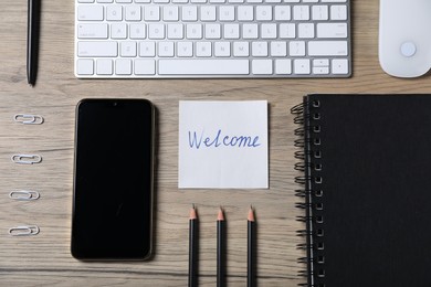 Photo of Paper note with word Welcome, smartphone, keyboard and stationery on wooden table, flat lay