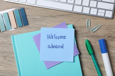Photo of Paper note with phrase Welcome Aboard, keyboard and stationery on wooden table, flat lay