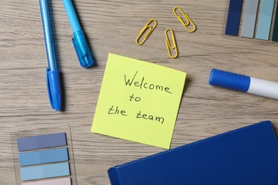 Photo of Paper note with phrase Welcome To The Team and stationery on wooden table, flat lay