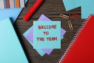 Photo of Paper note with phrase Welcome To The Team and stationery on wooden table, flat lay
