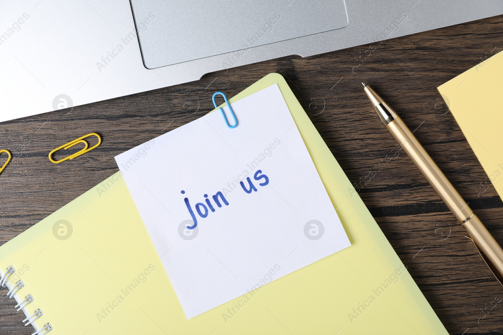 Photo of Paper note with phrase Join Us, laptop and stationery on wooden table, flat lay