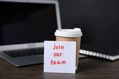 Photo of Paper note with phrase Join Our Team, cup of coffee, laptop and notebook on wooden table