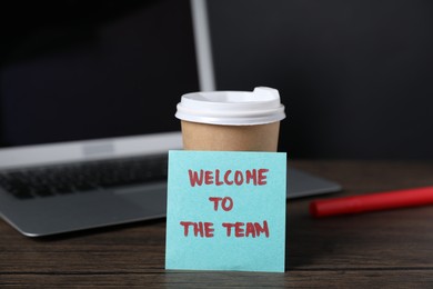 Photo of Paper note with phrase Welcome To The Team, cup of coffee, pen and laptop on wooden table