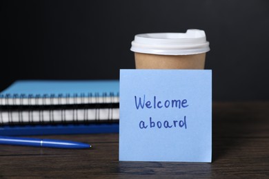 Photo of Paper note with phrase Welcome Aboard, cup of coffee and stationery on wooden table