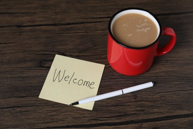 Photo of Paper note with word Welcome, cup of coffee and pen on wooden table, closeup