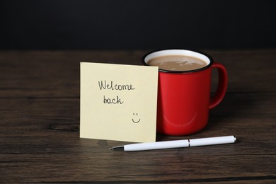 Photo of Paper note with phrase Welcome Back, pen and cup of coffee on wooden table