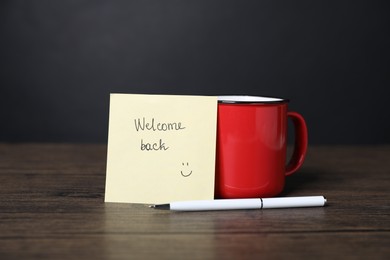 Photo of Paper note with phrase Welcome Back, pen and cup of coffee on wooden table
