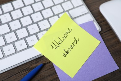 Photo of Paper note with phrase Welcome Aboard, pen and keyboard on wooden table, closeup