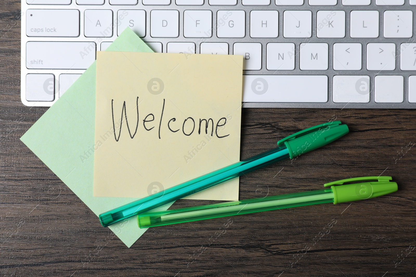 Photo of Paper note with word Welcome, pens and keyboard on wooden table, flat lay