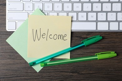 Photo of Paper note with word Welcome, pens and keyboard on wooden table, flat lay