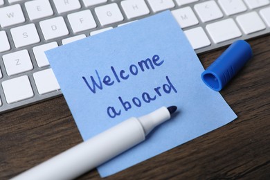 Photo of Paper note with phrase Welcome Aboard, marker and keyboard on wooden table, closeup