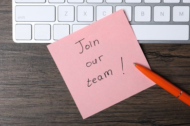 Photo of Paper note with phrase Join Our Team, pen and keyboard on wooden table, top view