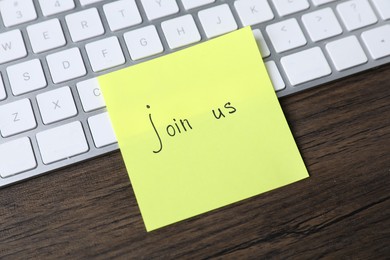 Photo of Paper note with phrase Join Us and keyboard on wooden table, closeup