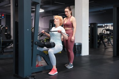 Photo of Senior woman training on lat pulldown machine supervised by professional trainer in gym