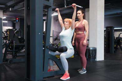 Photo of Senior woman training on lat pulldown machine supervised by professional trainer in gym