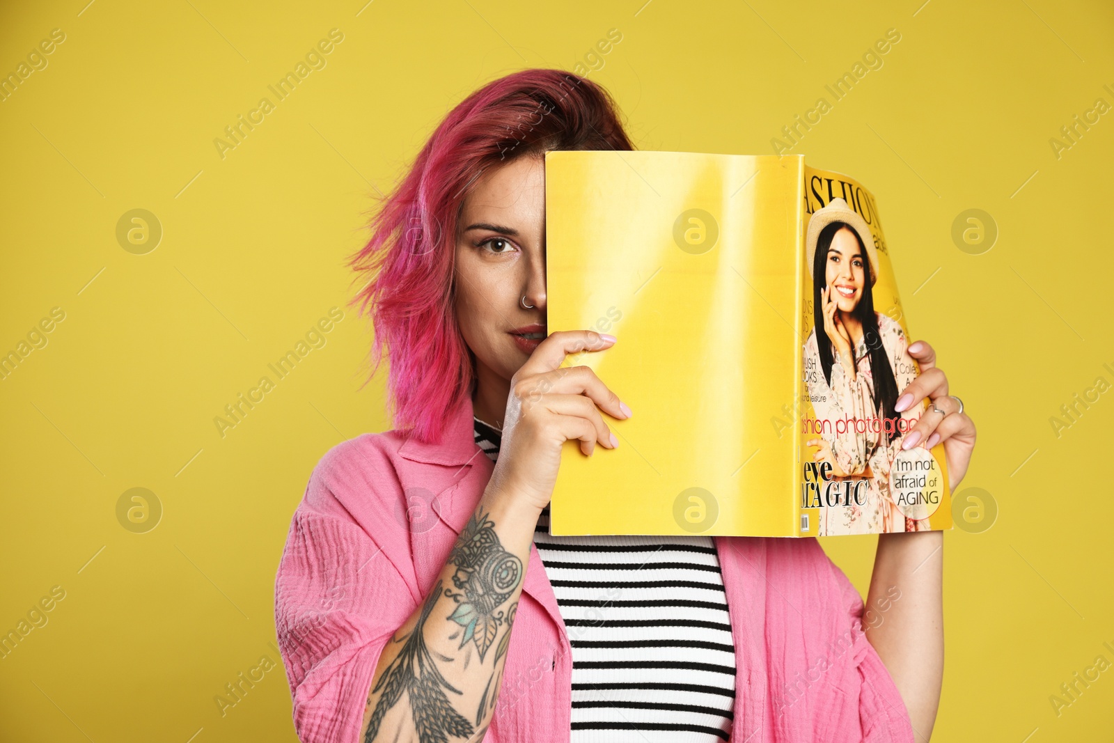 Beautiful woman with pink dyed hair covering eye with fashion magazine on yellow background Photo of Beautiful woman with pink dyed hair covering eye with fashion magazine on yellow background