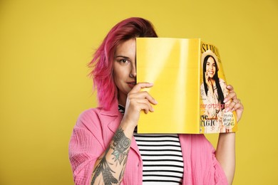 Photo of Beautiful woman with pink dyed hair covering eye with fashion magazine on yellow background