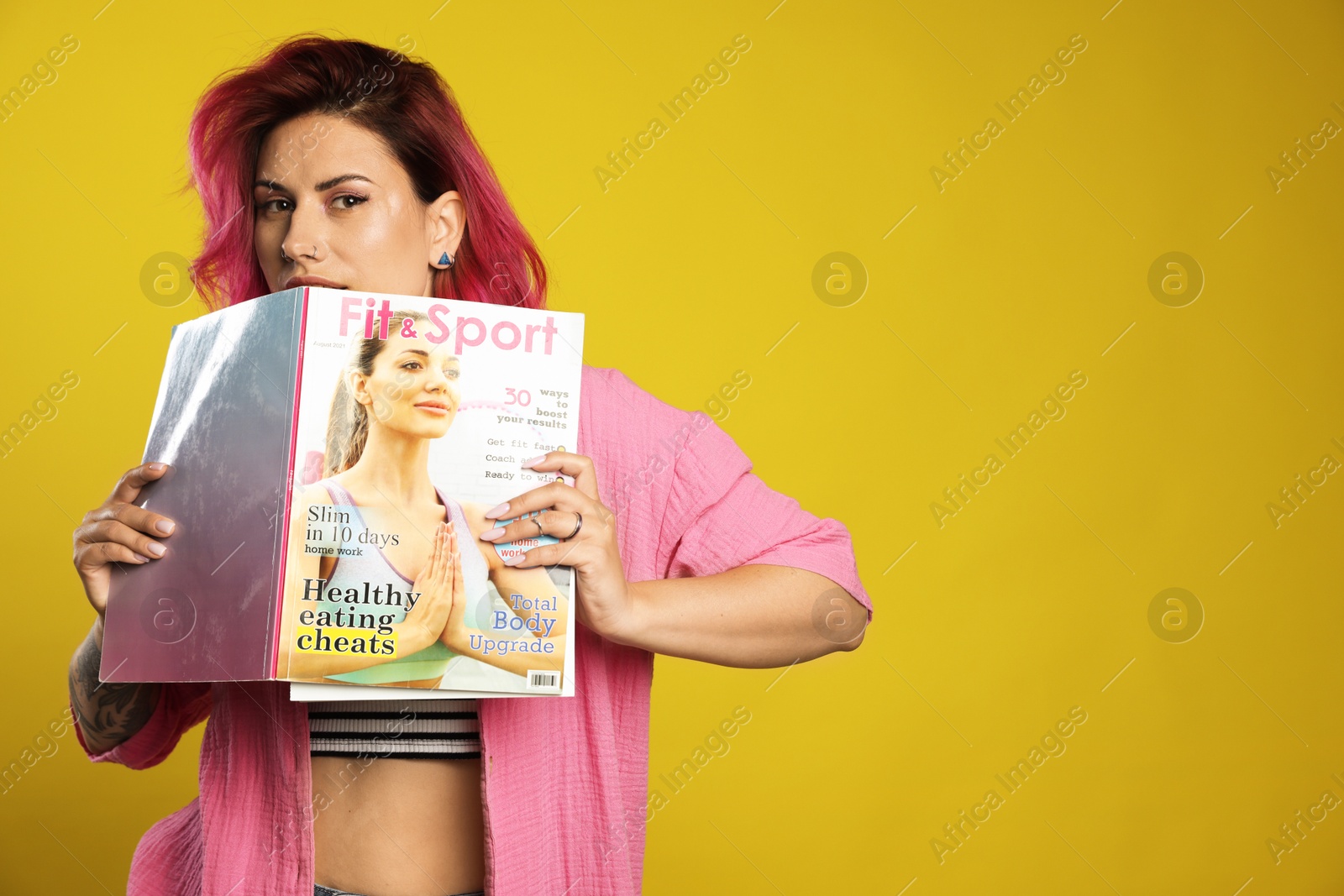 Beautiful woman with pink dyed hair holding sports magazine on yellow background, space for text Photo of Beautiful woman with pink dyed hair holding sports magazine on yellow background, space for text