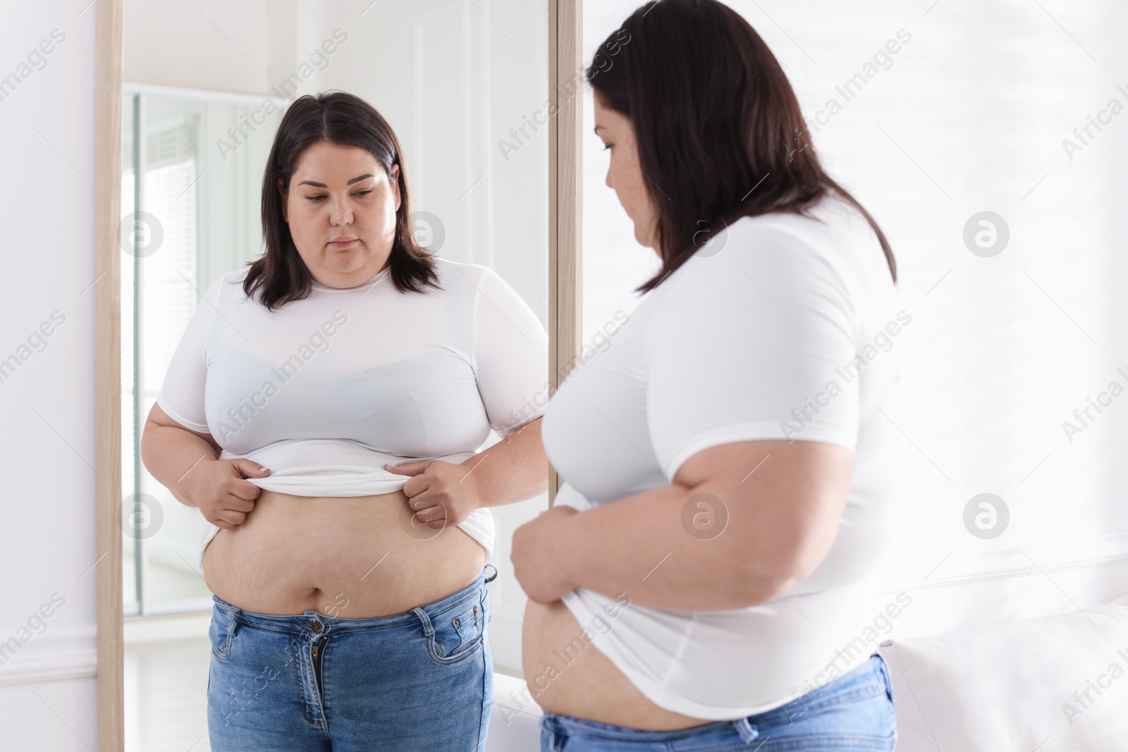 Woman with excessive belly fat near mirror indoors. Overweight problem Photo of Woman with excessive belly fat near mirror indoors. Overweight problem