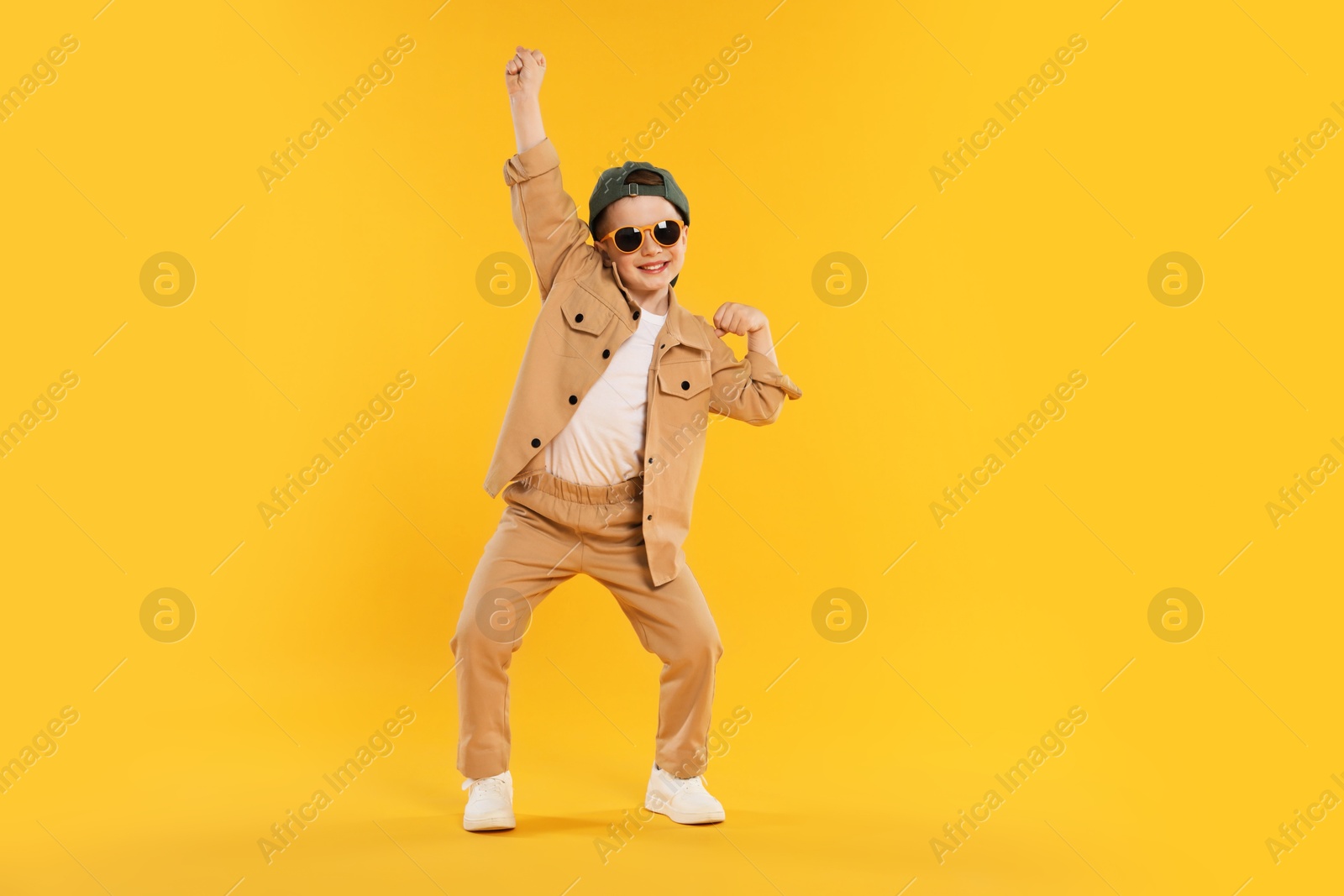 Photo of Happy little boy in sunglasses and baseball hat dancing on orange background