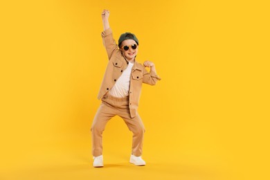 Photo of Happy little boy in sunglasses and baseball hat dancing on orange background
