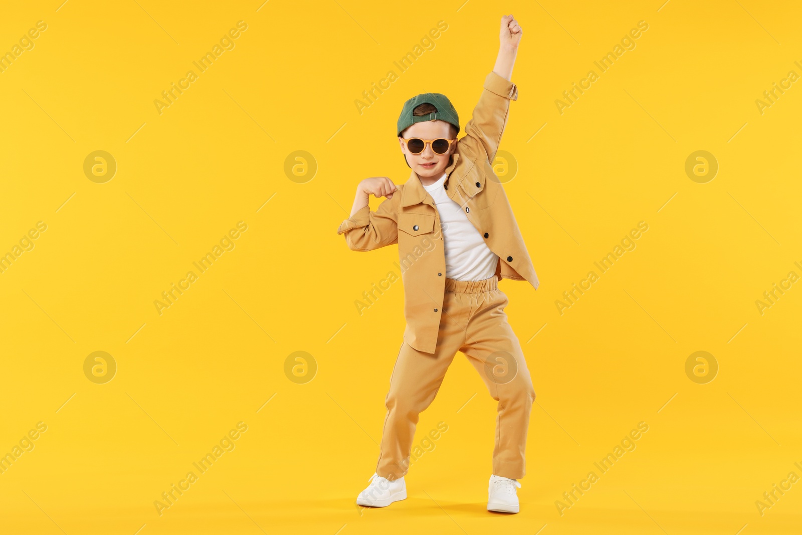 Photo of Cute little boy in sunglasses and baseball hat dancing on orange background. Space for text