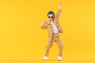 Photo of Cute little boy in sunglasses and baseball hat dancing on orange background. Space for text