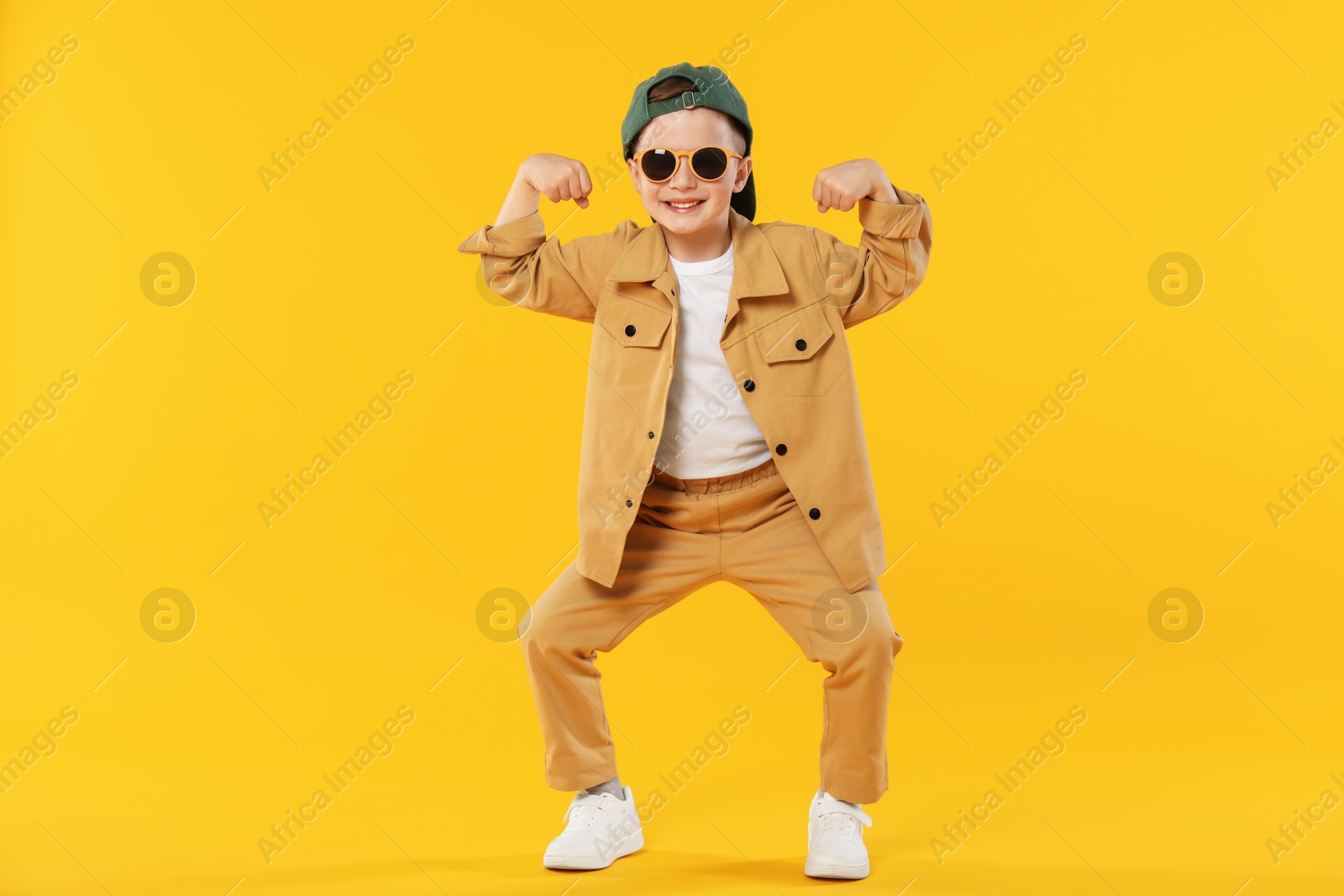 Happy little boy in sunglasses and baseball hat dancing on orange background Photo of Happy little boy in sunglasses and baseball hat dancing on orange background