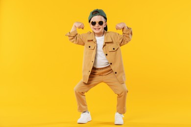 Photo of Happy little boy in sunglasses and baseball hat dancing on orange background
