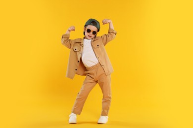 Photo of Cute little boy in sunglasses and baseball hat dancing on orange background