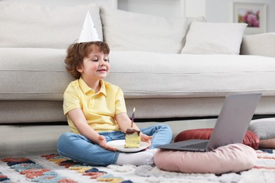 Photo of Birthday celebration. Little boy with piece of cake having video call via laptop at home