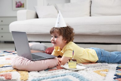 Photo of Birthday celebration. Little boy with piece of cake having video call via laptop at home