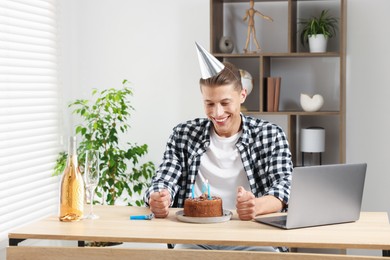 Photo of Birthday celebration. Man with cake and sparkling wine having video call via laptop at desk indoors