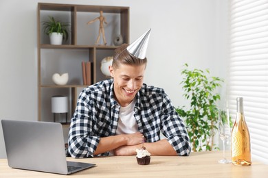 Photo of Birthday celebration. Man with cupcake and sparkling wine having video call via laptop at table indoors