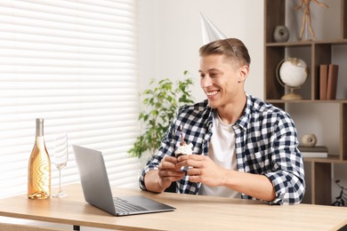 Photo of Birthday celebration. Man with cupcake and sparkling wine having video call via laptop at table indoors
