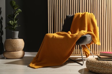 Photo of Comfortable armchair with orange woolen blanket, houseplant and books indoors