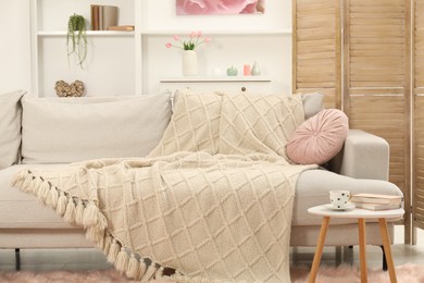 Photo of Comfortable sofa with woolen blanket, pillow, books and cup on side table indoors