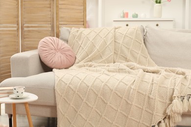 Photo of Comfortable sofa with woolen blanket, pillow, books and cup on side table indoors