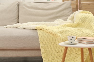 Photo of Comfortable sofa with woolen blanket, books and cup on side table indoors