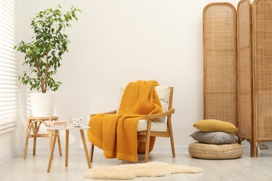 Photo of Comfortable armchair with orange woolen blanket, houseplant, books and cup on table indoors