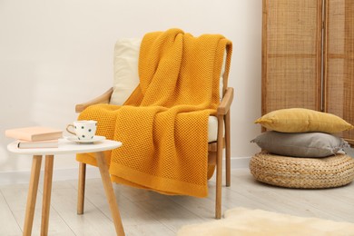 Photo of Comfortable armchair with orange woolen blanket, books and cup on table indoors