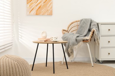 Photo of Wicker chair with gray woolen blanket, books, vase and cup on wooden table indoors