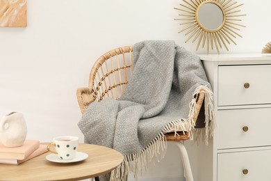 Photo of Wicker chair with gray woolen blanket, books, vase and cup on wooden table indoors