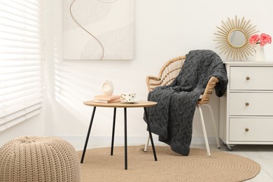 Photo of Wicker chair with gray woolen blanket, books, vase and cup on wooden table indoors
