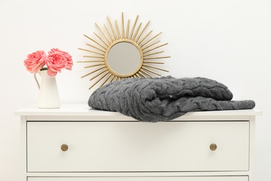 Photo of Gray woolen blanket, mirror and beautiful flowers in vase on white chest of drawers indoors