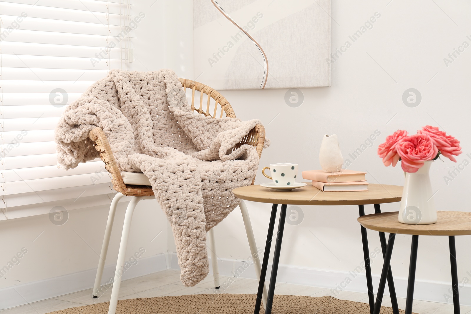 Wicker chair with woolen blanket, books, beautiful flowers in vase and cup on nesting tables indoors Photo of Wicker chair with woolen blanket, books, beautiful flowers in vase and cup on nesting tables indoors