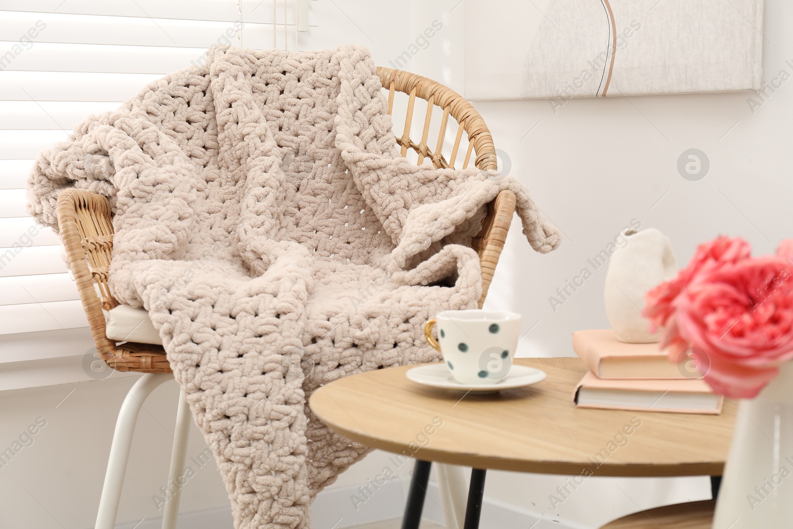 Wicker chair with woolen blanket, books, vase and cup on wooden table indoors Photo of Wicker chair with woolen blanket, books, vase and cup on wooden table indoors