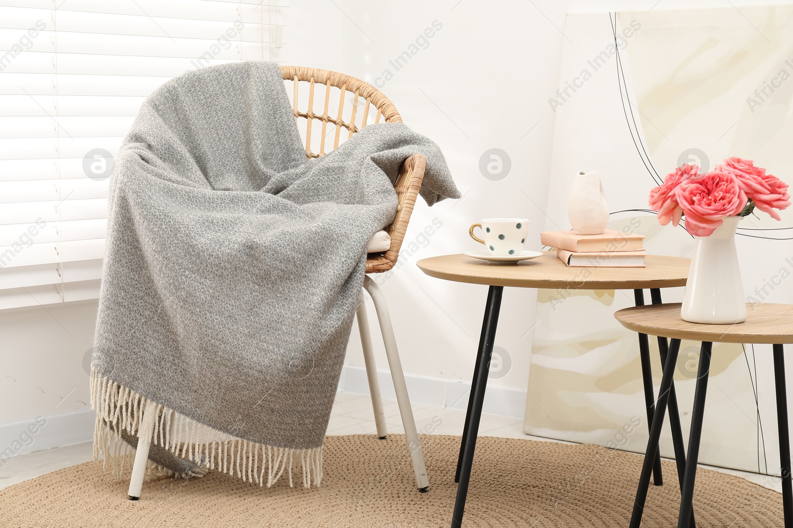 Wicker chair with gray woolen blanket, books, beautiful flowers in vase and cup on nesting tables indoors Photo of Wicker chair with gray woolen blanket, books, beautiful flowers in vase and cup on nesting tables indoors