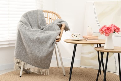 Photo of Wicker chair with gray woolen blanket, books, beautiful flowers in vase and cup on nesting tables indoors